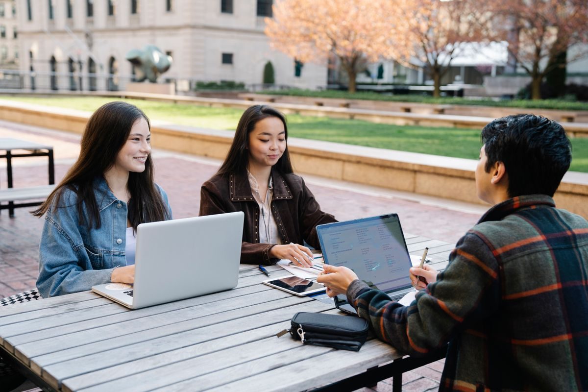 Three students at an outdoor table on campus working together on laptops